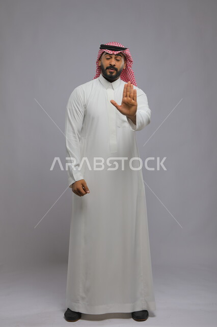 Prohibition of doing something, silence and stopping talking, portrait of a Saudi Gulf Arab man wearing the traditional thobe and shemagh, closing his eyes and raising his palm up, asking to lower the voice and threatening, full body portrait, gray background