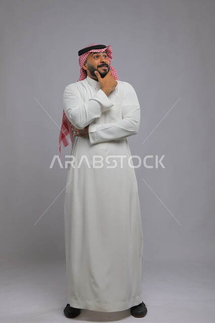 التأمل والتخطيط والتمعن بأمر محدد, بورتريه لشاب عربي خليجي سعودي يرتدي  الثوب والشماغ التقليدي يضع يديه على ذقنه بإيماءات التفكير, ايجاد الافكار والحلول, صورة لكامل طول الجسم, خلفية رمادية