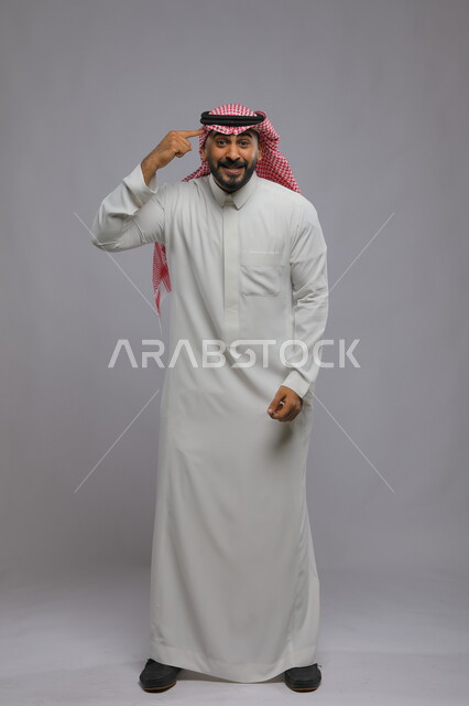Gestures of focus and planning, portrait of a young Saudi Gulf Arab man wearing a traditional dress and shemagh looking at the camera, the concept of thinking and finding a solution, full-length portrait of the body, gray background