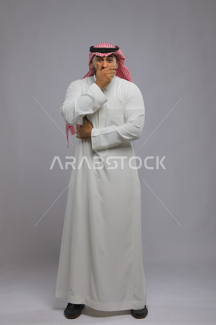 الشعور بالسعادة والفرحة الشديدة, المفاجأة والدهشة من شيء ما, مفهوم الانصات والتوقف عن الكلام, بورتريه لشاب عربي خليجي سعودي يرتدي الزي التقليدي يضع يديه على فمه بإيماءات الحياء والخجل, صورة الجسم بالطول الكامل, خلفية رمادية