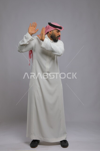 الاحتياط والقلق والابتعاد عن شيء ما, حماية النفس من ضرر خارجي, رفع اليدين بإيماءات التوقف وعدم الاقتراب, بورتريه لرجل عربي خليجي سعودي يرتدي الثوب التقليدي والشماغ يقف بحذر, صورة الجسم الكامل, خلفية رمادية