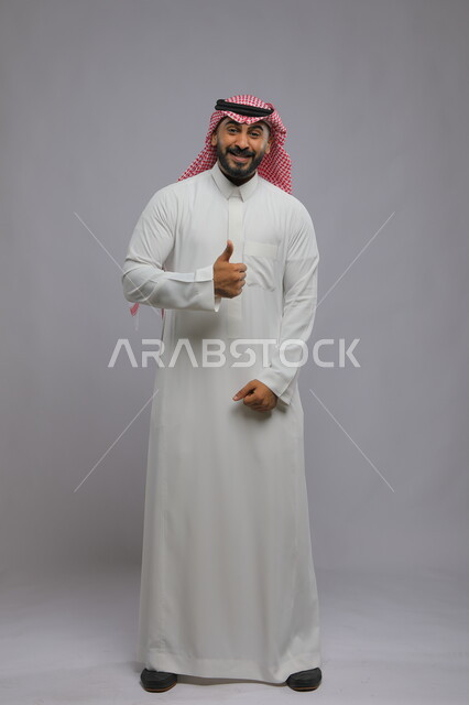 علامة الجودة والامتياز, بورتريه لرجل عربي خليجي سعودي يرتدي الثوب التقليدي والشماغ يرفع ابهامه بإيماءات الموافقة والقبول, الاعجاب والرضا لتحقيق أمر معين, صورة الجسم الكامل, خلفية رمادية