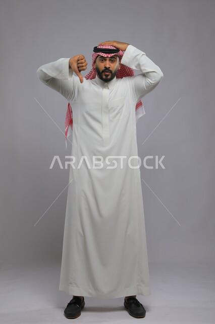 Anxiety, failure, and rejection of something, portrait of a Saudi Gulf Arab man wearing a traditional thobe and shemagh, raising his thumb and pointing downward in annoyance, lack of acceptance and satisfaction and preventing the use of a certain thing, full body portrait, gray background