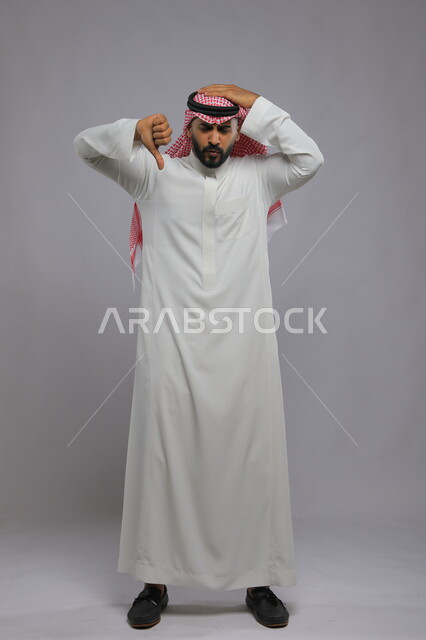 القلق والفشل ورفض شيء ما, بورتريه لرجل عربي خليجي سعودي يرتدي الثوب التقليدي والشماغ يرفع إبهامه ويشير للأسفل بانزعاج, عدم القبول والرضا ومنع استخدام شيء معين, صورة الجسم بالكامل, خلفية رمادية