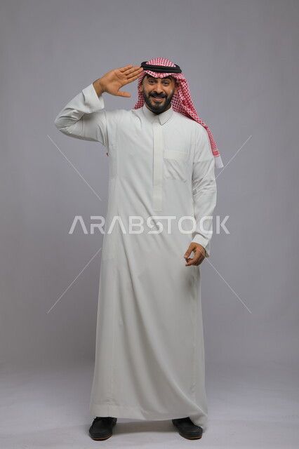 إلقاء التحية والسلام, التعبير عن الترحيب بسرور وبشاشة, بورتريه لرجل عربي خليجي سعودي يرتدي الثوب والشماغ التقليدي يرفع يديه على رأسه ويقف باستقامة, صورة الطول الكامل للجسم, خلفية رمادية
