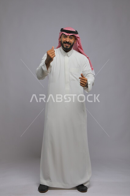 مفهوم الحب والإعجاب, ايماءات حركة القلب بالاصبعين, بورتريه لرجل عربي خليجي سعودي يرتدي الثوب والشماغ التقليدي يقف لطلب الأموال الكثيرة, السرور بالصفقات الناجحة والنمو الاقتصادي والمالي, صورة الجسم بالطول الكامل, خلفية رمادية