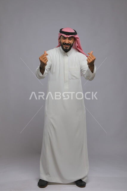 مفهوم الحب والإعجاب, ايماءات حركة القلب بالاصبعين, بورتريه لرجل عربي خليجي سعودي يرتدي الثوب والشماغ التقليدي يقف لطلب الأموال الكثيرة, السرور بالصفقات الناجحة والنمو الاقتصادي والمالي, صورة الجسم بالطول الكامل, خلفية رمادية