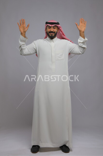 طلب الانقاذ والمساعدة, اللعب والانقياد والاستسلام, بورتريه لرجل عربي خليجي سعودي يرتدي الثوب التقليدي والشماغ يرفع يديه للأعلى بإيماءات الفرح والسعادة, صورة الجسم بالكامل, خلفية رمادية