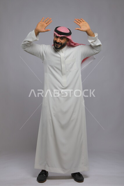 طلب الانقاذ والمساعدة, اللعب والانقياد والاستسلام, بورتريه لرجل عربي خليجي سعودي يرتدي الثوب التقليدي والشماغ يرفع يديه للأعلى بإيماءات الفرح والسعادة, صورة الجسم بالكامل, خلفية رمادية