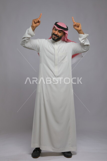 الاستقامة بالوقوف والنظر إلى الكاميرا, بورتريه لرجل عربي خليجي سعودي مبتسم يرتدي الثوب التقليدي والشماغ يرفع اصابعه  للأعلى بإيماءات التوضيح, ايماءات الاستفسار والاشارة إلى شيء ما, صورة الطول الكامل للجسم, خلفية رمادية