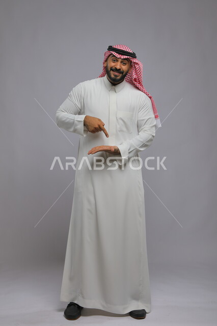 النظر إلى الكاميرا بإيماءات السرور, بورتريه لرجل عربي خليجي سعودي مبتسم يرتدي الثوب التقليدي والشماغ يفتح كفه للأعلى ويشير إلى عنصر ما, صورة لكامل الجسم, خلفية رمادية