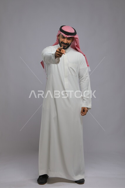 ايماءات التأكيد وتحديد الشيء المقابل, الإشارة الى شخص ما, بورتريه لرجل عربي خليجي سعودي يرتدي الثوب التقليدي والشماغ يرفع اصبعه السبابة للأمام, صورة لكامل الجسم, خلفية رمادية