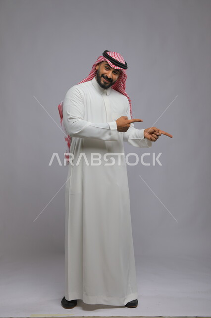البشاشة والسرور وتحديد شيء معين, الإشارة إلى مكان ما والنظر إلى الكاميرا, بورتريه لرجل عربي خليجي سعودي يرتدي الثوب التقليدي والشماغ يرفع إصبعه السبابة للاختيار, صورة الجسم بالكامل, خلفية رمادية