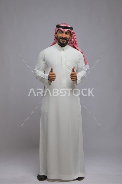 نظرات السرور والشعور بالرضى, بورتريه لرجل عربي خليجي سعودي يرتدي الثوب والشماغ التقليدي يرفع إبهامه للأعلى بعلامة الجودة, الوقوف باستقامة والنظر إلى الكاميرا بإيماءات التفاعل والموافقة, صورة الجسم بالطول الكامل,  خلفية رمادية