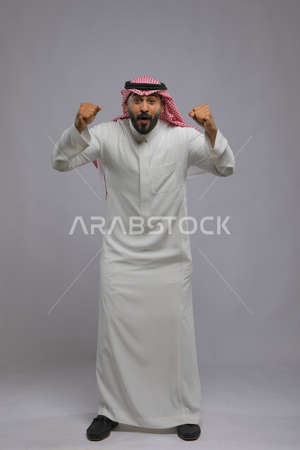 الاندماج بتشجيع كرة القدم, إيماءات الفوز والانتصار, بورتريه لرجل عربي خليجي سعودي مبتسم يرتدي الشماغ والثوب التقليدي يرفع يديه بتفاعل وحماس, صورة بالطول الكامل للجسم, خلفية رمادية
