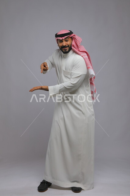Looking at the camera and identifying a specific thing with gestures of pleasure, portrait of a smiling Saudi Gulf Arab man wearing a traditional thobe and shemagh, opening his palm up and pointing to an item, full-length portrait of the body, gray background