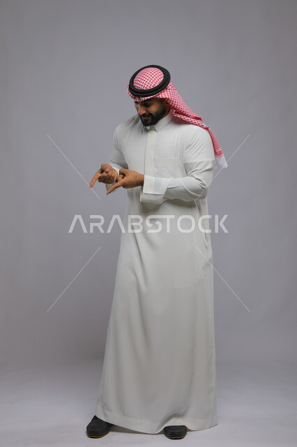 الإشارة وتحديد شيء ما, بورتريه لرجل عربي خليجي سعودي يرتدي الثوب التقليدي والشماغ يرفع أصابعه ويشير إلى هذا المكان بإيماءات الدهشة والتأكيد, صورة الجسم بالطول الكامل, خلفية رمادية