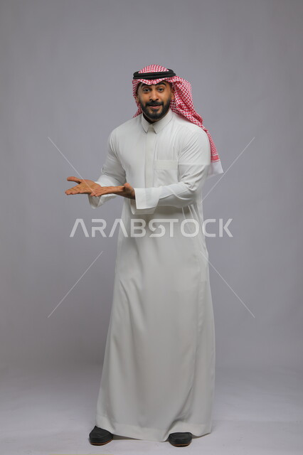 Standing straight and looking at the camera, portrait of a Saudi Gulf Arab man wearing a traditional thobe and shemagh, opening his palms upwards with gestures of holding an item, pointing and pointing with enthusiasm and amazement, full-length portrait of the body, gray background