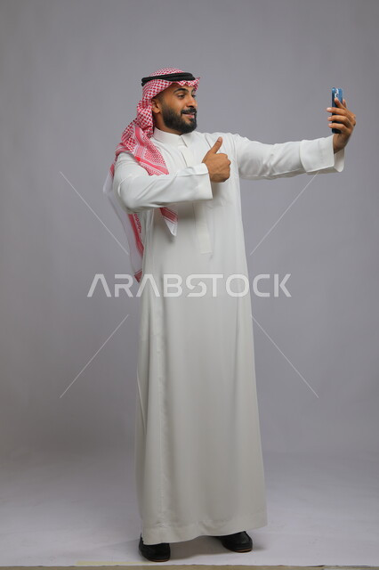 استخدام الهاتف المحمول لالتقاط الصور التذكارية،  بورتريه لرجل عربي سعودي خليجي مبتسم يرتدي الثوب والشماغ التقليدي يرفع اصبعه نحو الأعلى بعلامة الجودة,الشعور بالرضا والقبول، إجراء مكالمات فيديو بإيماءات الفرح والسرور, صورة الجسم بالطول الكامل, خلفية رمادية.