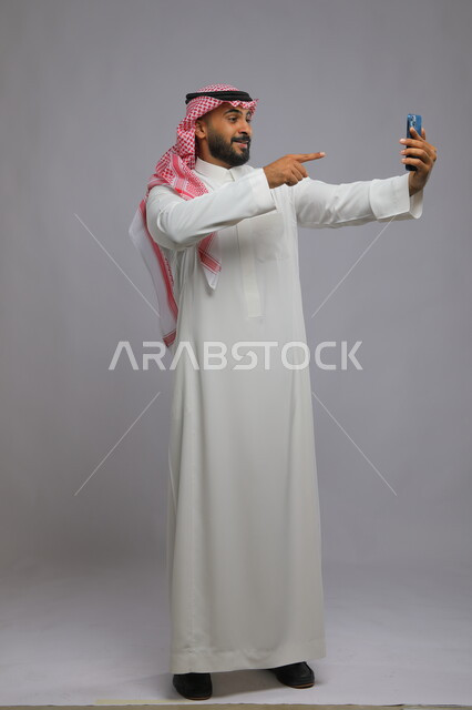 استخدام التقنيات الحديثة لالتقاط الصور التذكارية، إجراء مكالمات فيديو بإيماءات الفرح والسرور, بورتريه لشاب عربي سعودي خليجي مبتسم يرتدي الثوب والشماغ التقليدي يشير إلى شاشة الهاتف المحمول, التواصل مع الأهل والأصدقاء, صورة الجسم بالطول الكامل, خلفية رمادية.