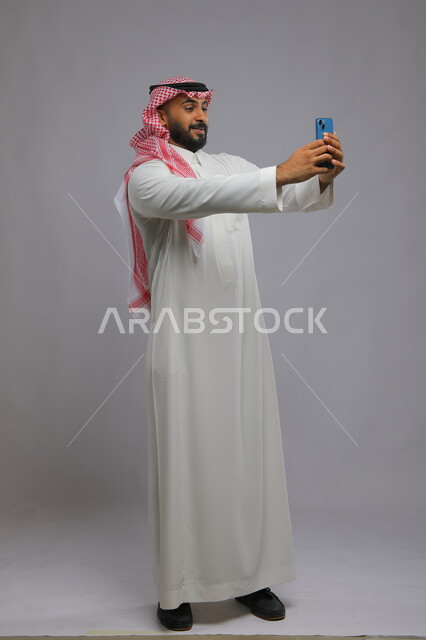 Taking souvenir photos via mobile phone and modern technology, a portrait of a Saudi Gulf Arab man wearing the traditional thobe and shemagh, looking using a mobile phone, live broadcast on social media, full body photo, gray background