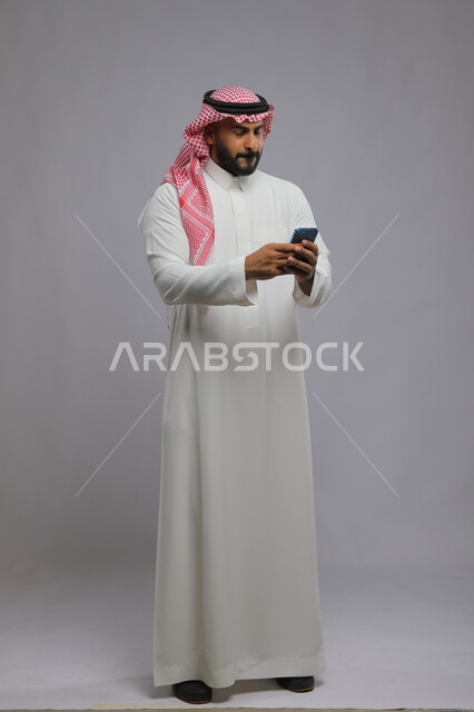 الشعور بالخوف والقلق لانتظار أمر ما, بورتريه لشاب عربي سعودي خليجي يرتدي الثوب والشماغ التقليدي يستخدم الهاتف المحمول ويغمض عينيه بإيماءات القلق, فقدان بيانات الدخول, صورة الجسم بالكامل, خلفية رمادية.