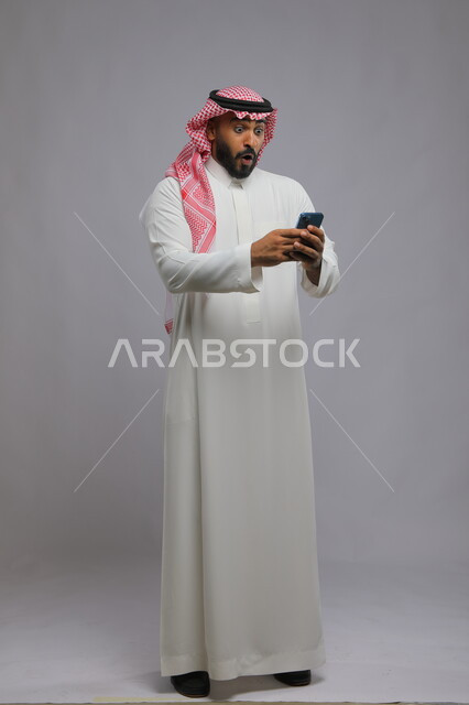  الشعور بالحماس للنجاح وتحقيق الأهداف, بورتريه لشاب عربي سعودي خليجي يرتدي الثوب والشماغ التقليدي يستخدم الهاتف المحمول, إيماءات الاندهاش والذهول, ميزات تقنية حديثة ومتطورة, صورة الجسم بالكامل, خلفية رمادية.