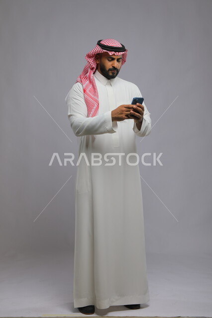 الشعور بالانزعاج لمعرفة الأخبار السيئة, إيماءات الغضب والعبوس, بورتريه لشاب عربي سعودي خليجي يرتدي الثوب والشماغ التقليدي يستخدم الهاتف المحمول, دردشات ومحادثات عبر الانترنت, صورة الجسم بالكامل, خلفية رمادية.