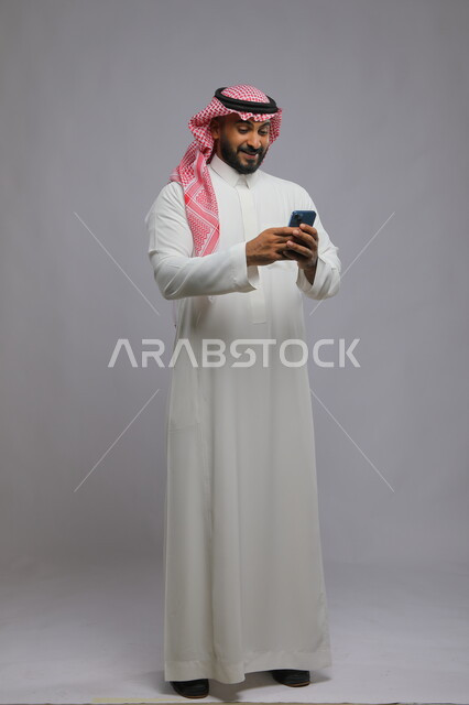 التواصل مع الأهل والأصدقاء, إجراء مكالمات فيديو بإيماءات الفرح والسرور, بورتريه لشاب عربي سعودي خليجي يرتدي الثوب والشماغ التقليدي يستخدم الهاتف المحمول, تصفح تطبيقات وبرامج التواصل الاجتماعي, صورة الجسم بالطول الكامل, خلفية رمادية.
