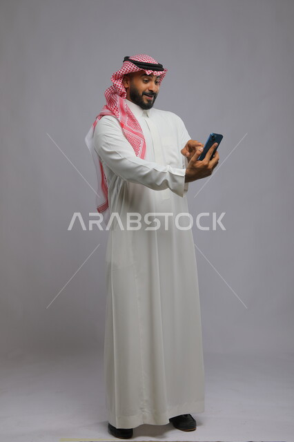 Making video calls with gestures of joy and happiness, portrait of a Saudi Gulf Arab young man wearing the traditional thobe and shemagh pointing to the mobile phone screen, communicating with family and friends, full-length body photo, gray background