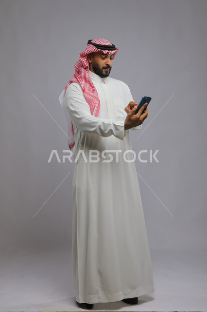 Pressing and clicking using the sense of touch, portrait of a smiling Saudi Arabian Gulf man wearing a traditional thobe and shemagh holding a mobile phone, browsing social media applications and programmes, modern and advanced technologies, full-length body image, gray background
