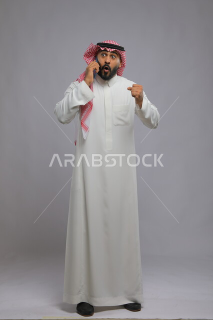 إيماءات الاندهاش والذهول, بورتريه لشاب عربي سعودي خليجي يرتدي الثوب والشماغ التقليدي يستخدم الهاتف المحمول, التواصل مع الأهل والأصدقاء, الشعور بالحماس لسماع الأخبار السارة, صورة الجسم بالكامل, خلفية رمادية.