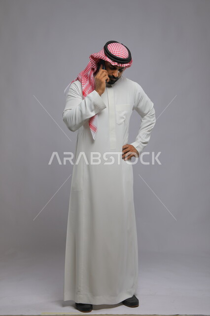 الشعور بالحيرة والقلق, بورتريه لرجل عربي سعودي خليجي يرتدي الثوب والشماغ التقليدي يقف ويضع يده على خصره يستخدم الهاتف المحمول, التواصل مع الأهل والأصدقاء للاطمئنان, مناقشة أمر ما, صورة الجسم بالكامل, خلفية رمادية.