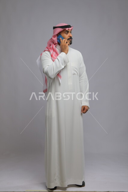 إجراء مكالمات هاتفية لمناقشة أمر ما, بورتريه لشاب عربي خليجي سعودي يرتدي الثوب والشماغ التقليدي يستخدم الهاتف المحمول, التواصل مع زملاء العمل, إيماءات القلق بشأن أمر ما, صورة لكامل الجسم, خلفية رمادية.