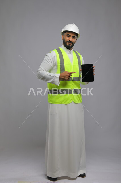 تسهيل القيام بمهام العمل اليومية عبرالتابلت والتقنيات الحديثة، الإشارة إلى شاشة سوداء فارغة, بورتريه لمهندس معماري عربي خليجي سعودي مبتسم يرتدي الخوذة وسترة الحماية ينظر إلى الكاميرا، صورة الجسم بالطول الكامل, خلفية رمادية.