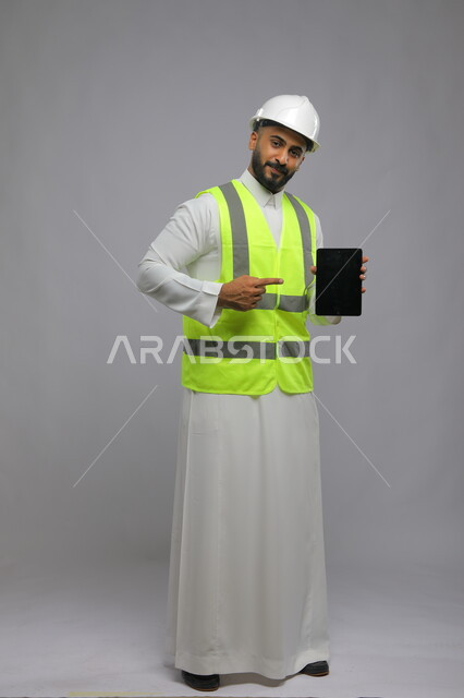 Pointing to a blank black screen, using a tablet and modern technologies to facilitate daily work tasks, portrait of a smiling Saudi Arabian Gulf architect wearing a helmet and protective vest looking at the camera, full-length body portrait, gray background