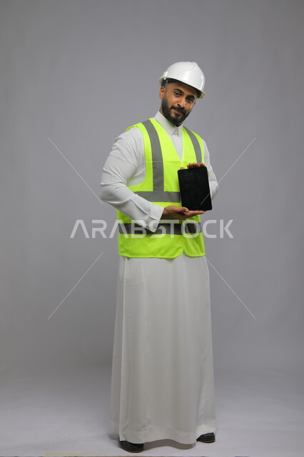 عرض شاشة سوداء فارغة, استخدام التابلت والتقنيات الحديثة لتسهيل القيام بمهام العمل اليومية، بورتريه لمهندس معماري عربي خليجي سعودي مبتسم يرتدي الخوذة وسترة الحماية ينظر إلى الكاميرا، صورة الجسم بالطول الكامل, خلفية رمادية.