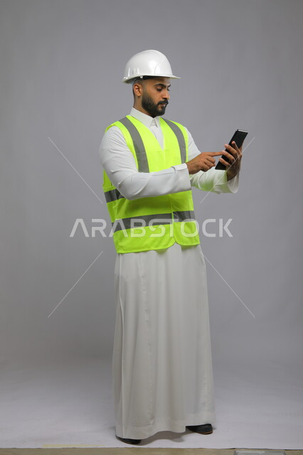 Using the tablet and modern technology to facilitate daily work tasks, reviewing plans and engineering projects, portrait of a Saudi Arabian Gulf civil engineer wearing a helmet and protective vest, using the tablet, focus and scrutiny gestures, full-length body image, gray background