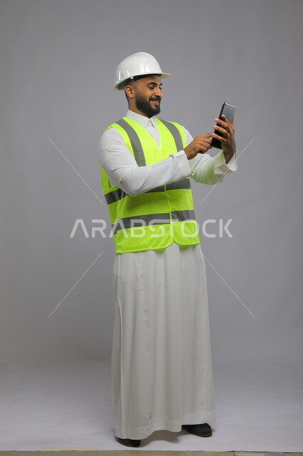 Integrating work with technology and technology in practical life, viewing new engineering designs via the tablet, portrait of a smiling Saudi Gulf Arab architect wearing a helmet and protective vest using a tablet, full-length body image, gray background