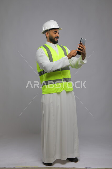 النمو والتطور العمراني في السعودية, دمج العمل مع التقنية و التكنولوجيا، الاطلاع على تصاميم هندسية جديدة عبر الجهاز اللوحي، بورتريه لمهندس معماري عربي خليجي سعودي مبتسم يرتدي الخوذة وسترة الحماية يستخدم التابليت، صورة الجسم بالطول الكامل, خلفية رمادية.