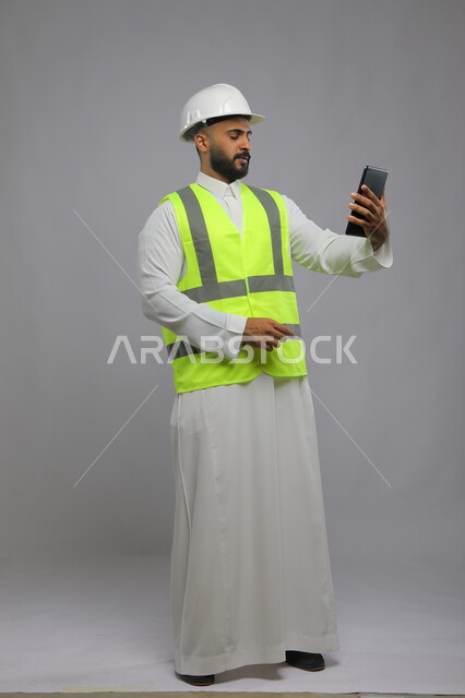 الاطلاع على تصاميم هندسية جديدة عبر التابليت، دمج العمل مع التقنية و التكنولوجيا، بورتريه لمهندس مدني عربي خليجي سعودي يرتدي الخوذة وسترة الحماية يغمض عينيه يستخدم الجهاز اللوحي، إيماءات التفكير والتخطيط لشيء ما, صورة الجسم بالطول الكامل, خلفية رمادية.