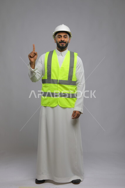الاشارة بأصبع السبابه الى الأعلى، متابعة سير المشاريع الإعمارية، تطور المباني على يد أبناء الوطن ،مفهوم الانشاءات المعمارية والهندسة، بورتريه لمهندس عربي خليجي سعودي مبتسم يرتدي الخوذة وسترة الحماية و ينظر إلى الكاميرا، صورة الجسم بالطول الكامل، خلفية رمادية