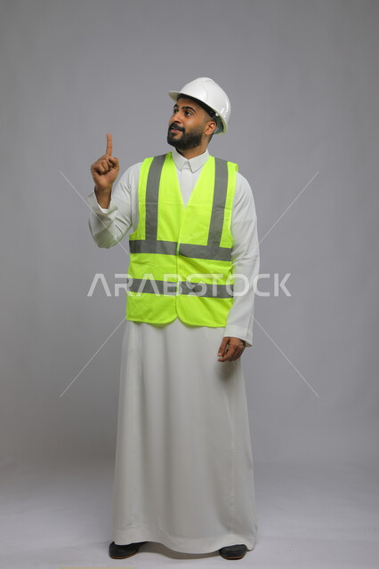 متابعة سير المشاريع الإعمارية، وظائف ومهن سعودية، تطور وتقدم المباني على يد مهندسين سعوديين ،مفهوم الانشاءات المعمارية والهندسة، بورتريه لمهندس عربي خليجي سعودي مبتسم يرتدي الخوذة وسترة الحماية يشير بأصبعه إلى الأعلى ، صورة الجسم بالطول الكامل، خلفية رمادية