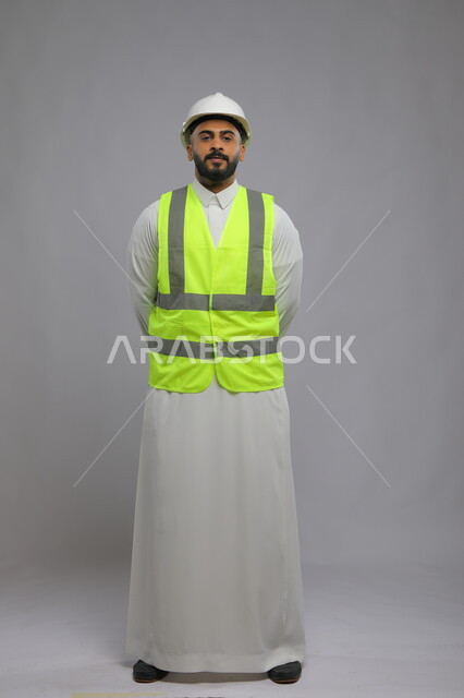 الإعمار بسواعد أبناء الوطن ،العمل في القطاع الهندسي، مفهوم الهندسة والانشاءات المعمارية، بورتريه لمهندس عربي خليجي سعودي يرتدي الخوذه و سترة الحماية ينظر إلى الكاميرا ويضع يديه خلف ظهره، مهن ووظائف سعودية، صورة لكامل الجسم، خلفية رمادية