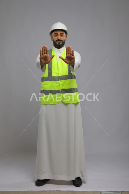 اشارات يدين تدل على الاحساس و اللمس ،تطور وتقدم المباني على يد أبناء الوطن ،مفهوم الانشاءات المعمارية والهندسة، بورتريه لمهندس عربي خليجي سعودي مبتسم يرتدي الخوذة وسترة الحماية يمد يده للأمام بإيماءات طلب التوقف والابتعاد، صورة الجسم بالطول الكامل، خلفية رمادية