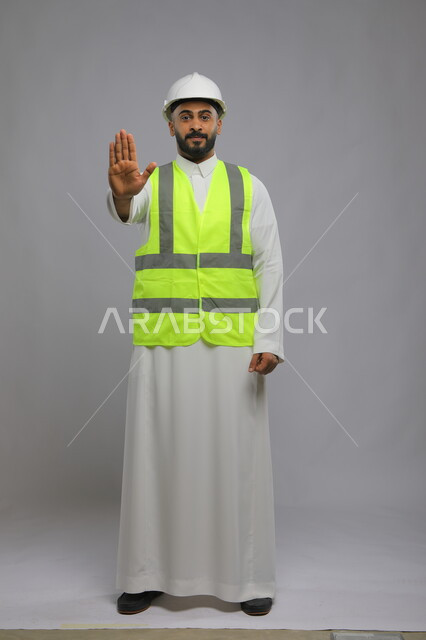 Gestures requesting to stop and move away, the development and progress of buildings at the hands of the people of the nation, the concept of architectural and engineering constructions, a portrait of a smiling Saudi Arabian Gulf engineer wearing a helmet and protection vest, extending his hand forward to refrain from doing something, full-length body portrait, gray background