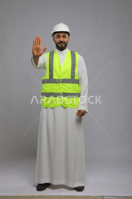 الامتناع عن فعل شيء ما، تطور وتقدم المباني على يد أبناء الوطن ،مفهوم الانشاءات المعمارية والهندسة، بورتريه لمهندس عربي خليجي سعودي مبتسم يرتدي الخوذة وسترة الحماية يمد يده للأمام بإيماءات التوقف والابتعاد، صورة الجسم بالطول الكامل، خلفية رمادية