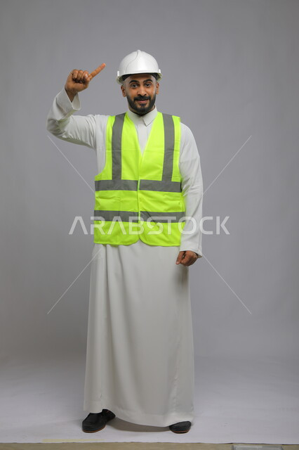 وظائف ومهن سعودية، تطور وتقدم المباني على يد مهندسين سعوديين ،مفهوم الانشاءات المعمارية والهندسة، بورتريه لمهندس عربي خليجي سعودي مبتسم يرتدي اللباس الخاص بالعمل يشير بأصبعه إلى خوذة الحماية، صورة الجسم بالطول الكامل ، خلفية رمادية