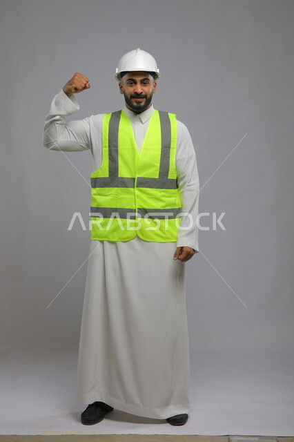 الفن في تقنية البناء والتصميم، الإنشاءات المعمارية في الأراضي السعودية ،متابعة سير المشاريع الإعمارية بجودة عالية، بورتريه لمهندس عربي خليجي سعودي مبتسم يرتدي الخوذه و سترة الحماية يرفع يده للأعلى بإيماءات النصر والنجاح، صورة الجسم بالكامل، خلفية رمادية
