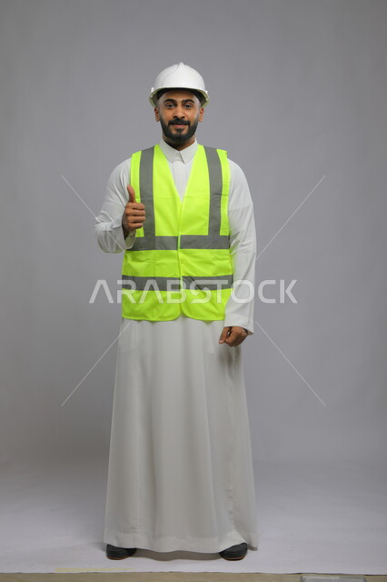 الإنشاءات المعمارية في الأراضي السعودية ،متابعة سير المشاريع الإعمارية بجودة عالية، الفن في تقنية التصميم والبناء، بورتريه لمهندس عربي خليجي سعودي مبتسم يرتدي الخوذه و سترة الحماية يرفع الابهام للأعلى بحركات الاعجاب والقبول، صورة الجسم بالكامل، خلفية رمادية